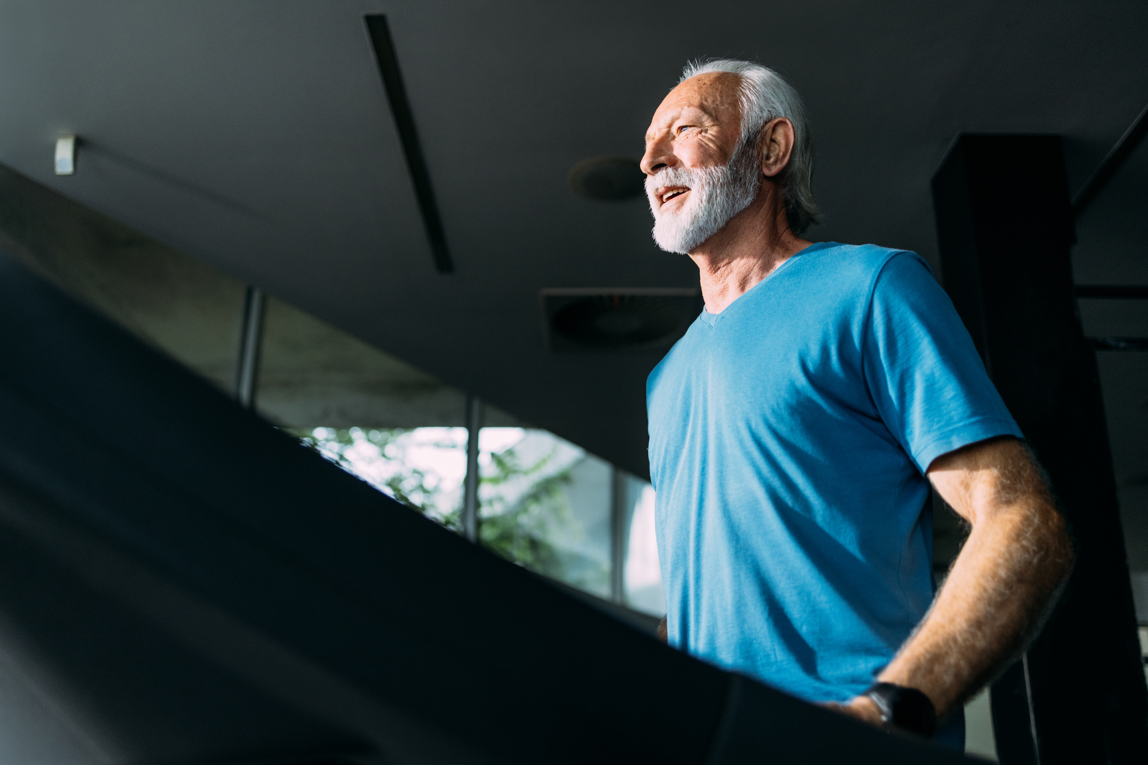 An older man with a white beard and mustache wearing a blue shirt is smiling while walking on a treadmill indoors