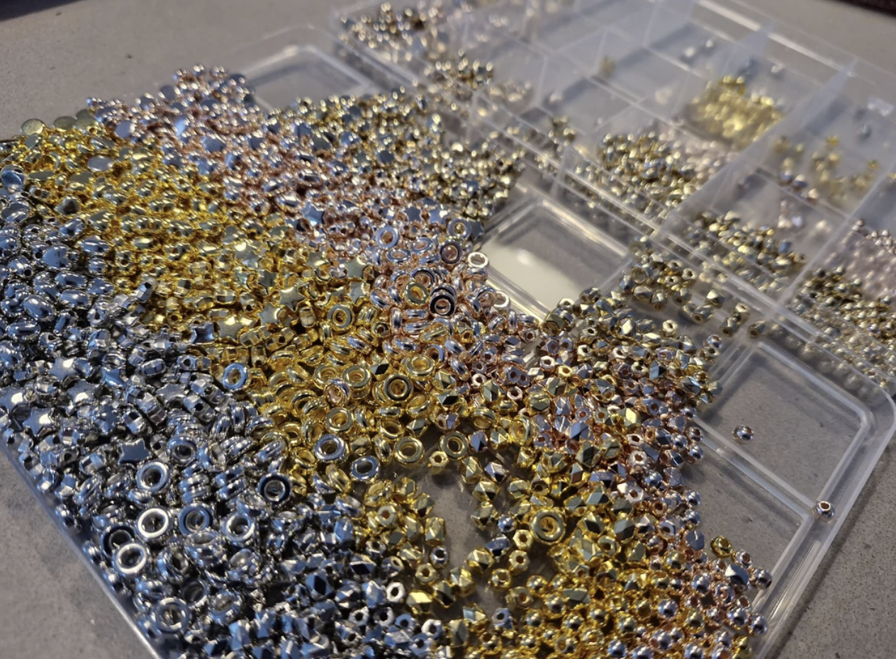 A large pile of small metallic beads in various shapes and sizes, with gold, silver, and rose gold finishes, arranged next to a clear plastic organizer