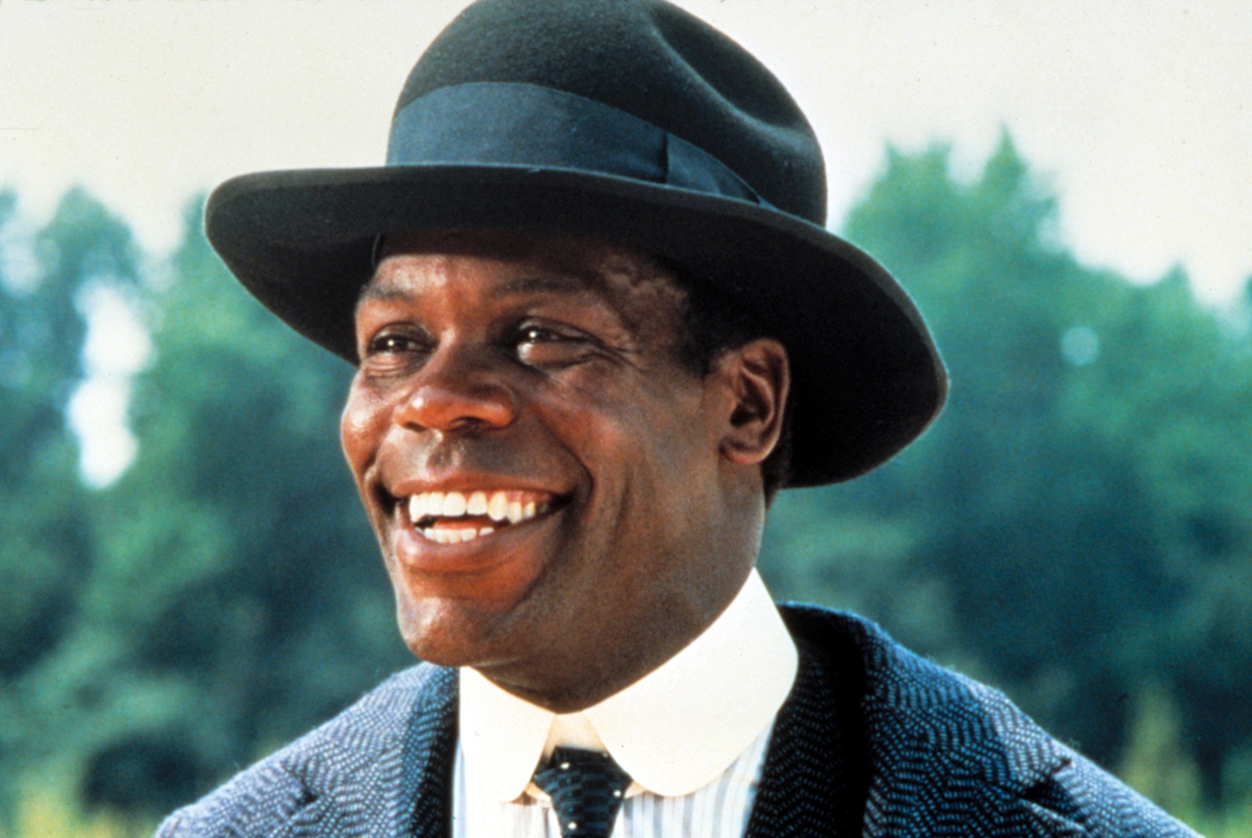 Danny Glover smiling, wearing a hat, a white shirt, a tie, and a textured jacket