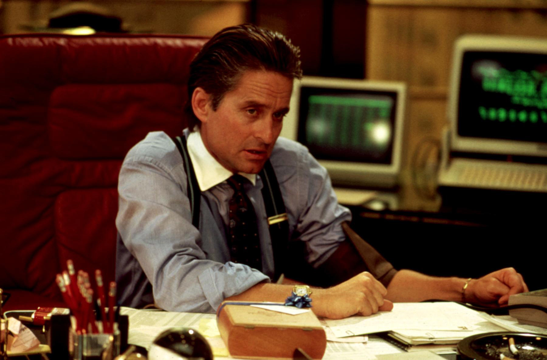 Michael Douglas, in a business suit with rolled-up sleeves, is seated at a cluttered desk with computers and papers in a scene from &quot;Wall Street&quot;