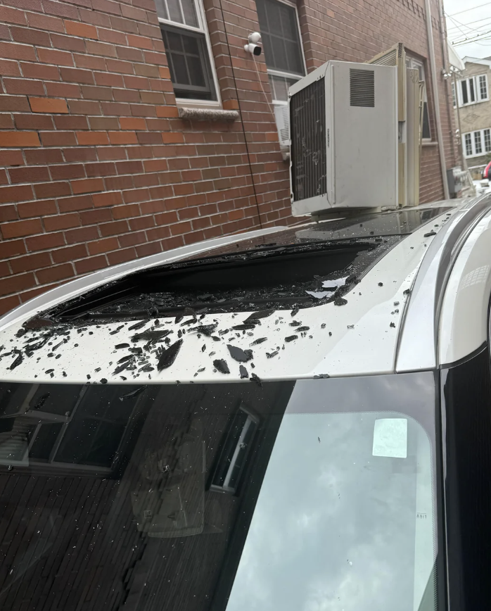 A car with a shattered sunroof parked next to a building with a window air conditioning unit above. Broken glass is scattered on the car roof