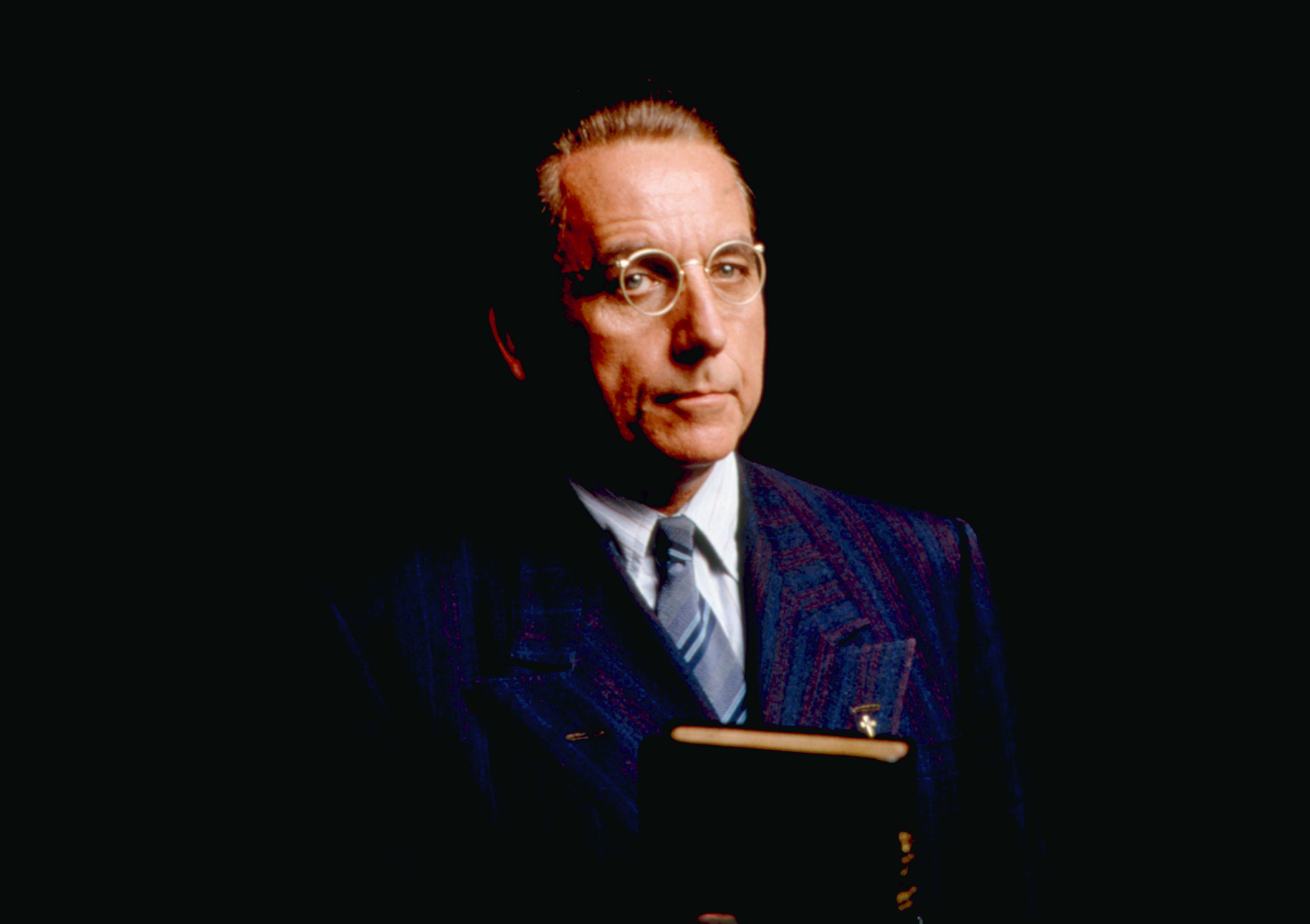 A man in formal attire, wearing a suit and tie, holds an object while looking ahead against a dark background