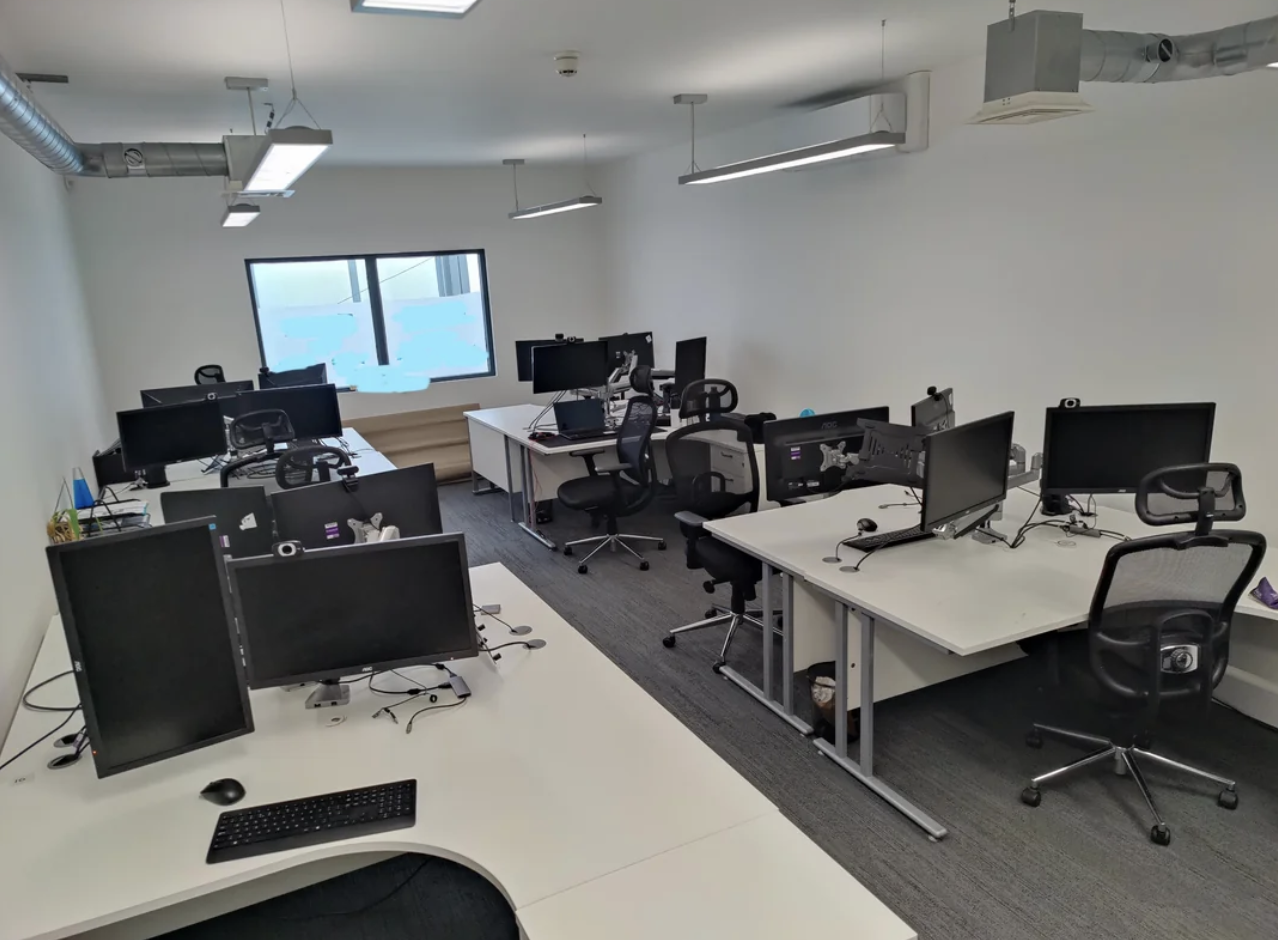 Empty modern office with multiple computer workstations, ergonomic chairs, and a window at the far end. No people are present