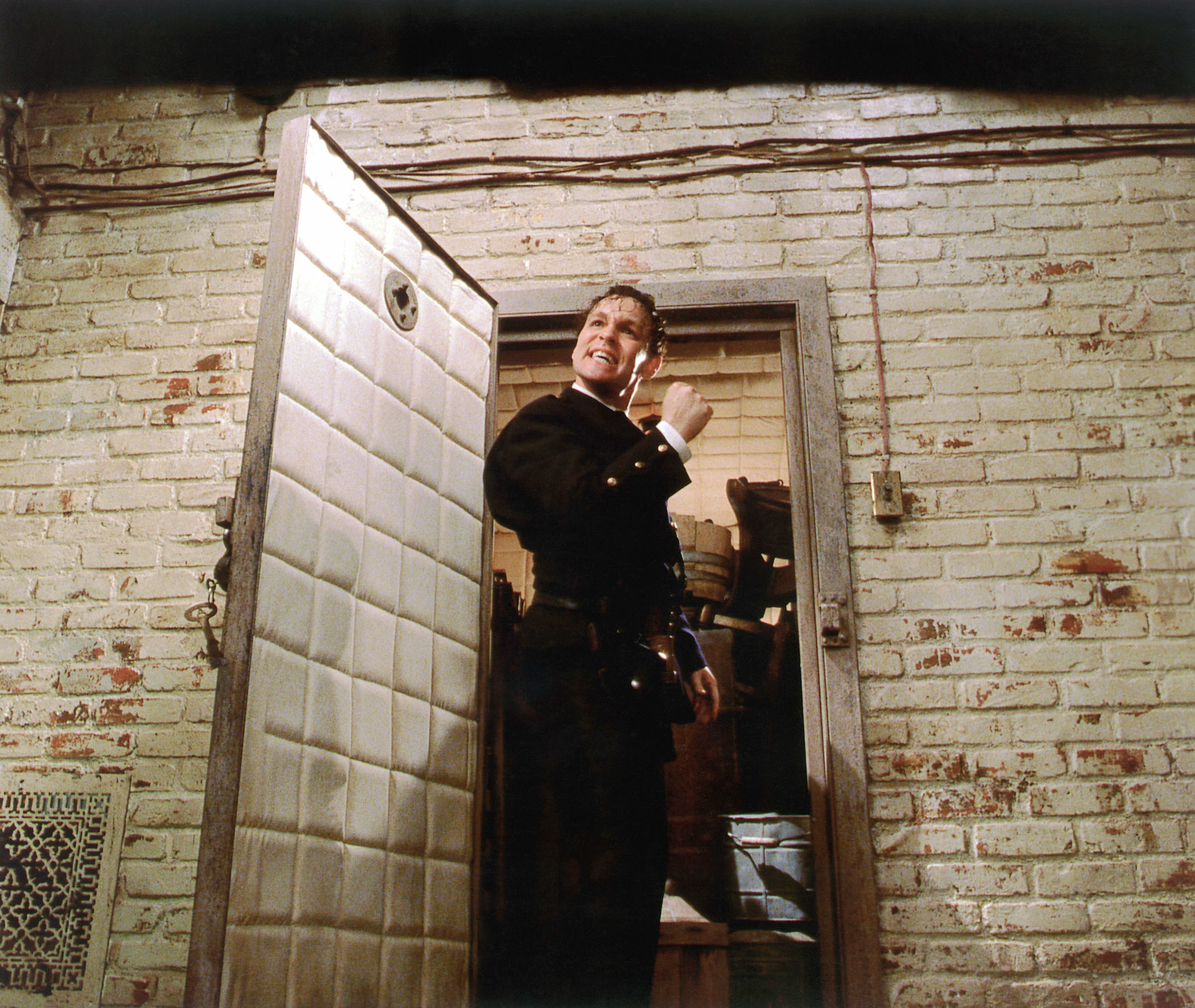 A man dressed in a dark uniform stands in a doorway of a brick building, looking upward with a smile and making a fist gesture