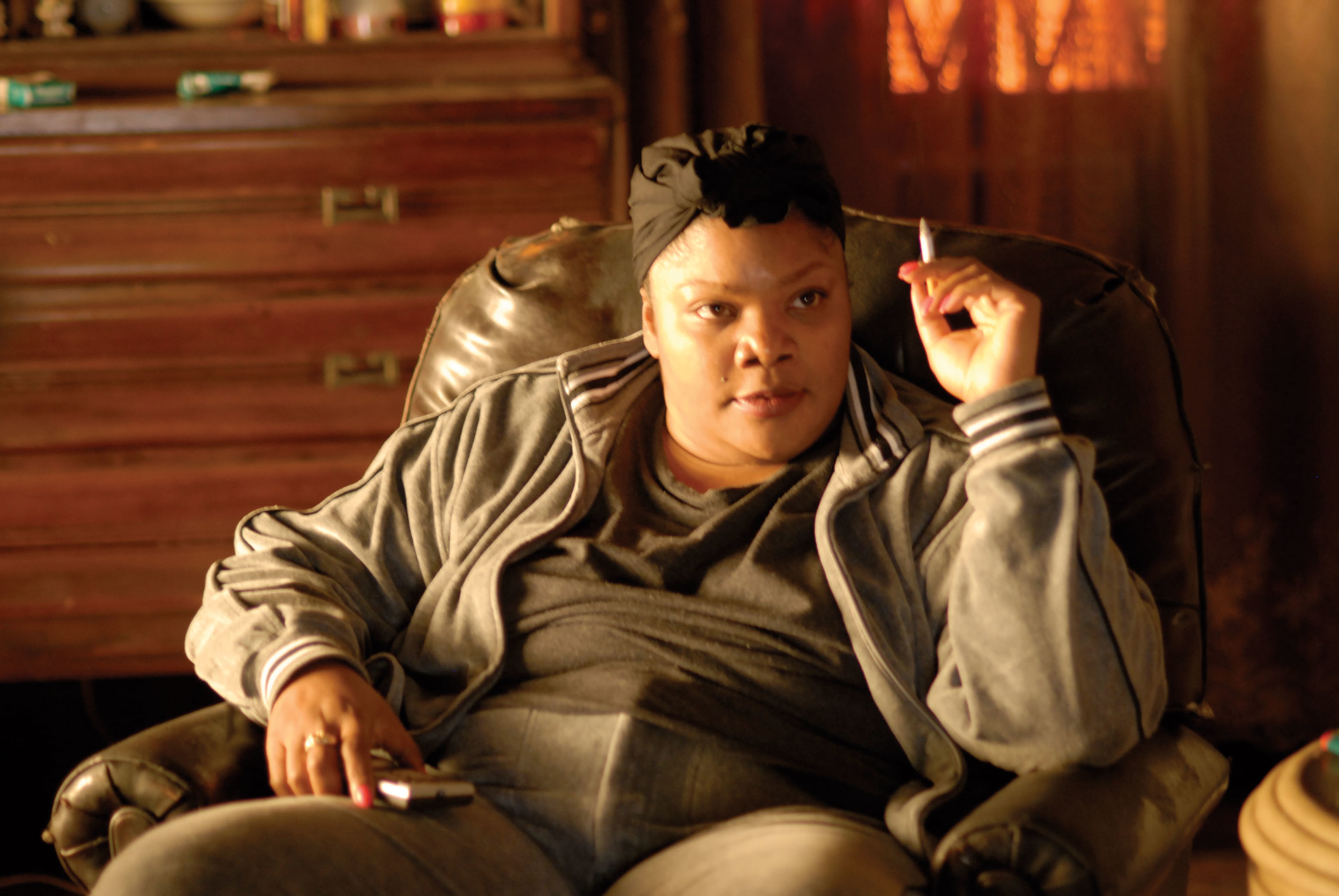 Mo&#x27;Nique in a casual outfit sits in a leather armchair, holding a cigarette in one hand and a TV remote in the other, appearing focused