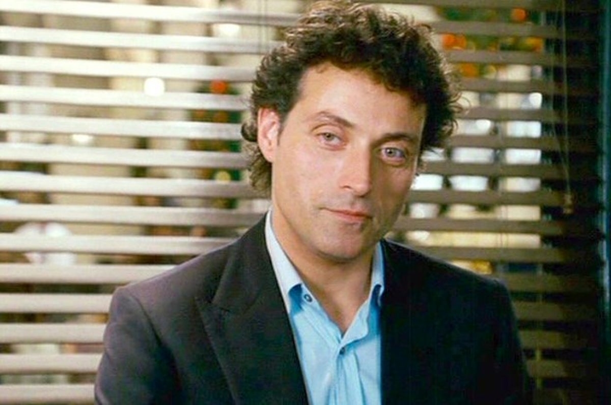 Rufus Sewell, in a suit with an open-collared shirt, poses in front of a window with blinds