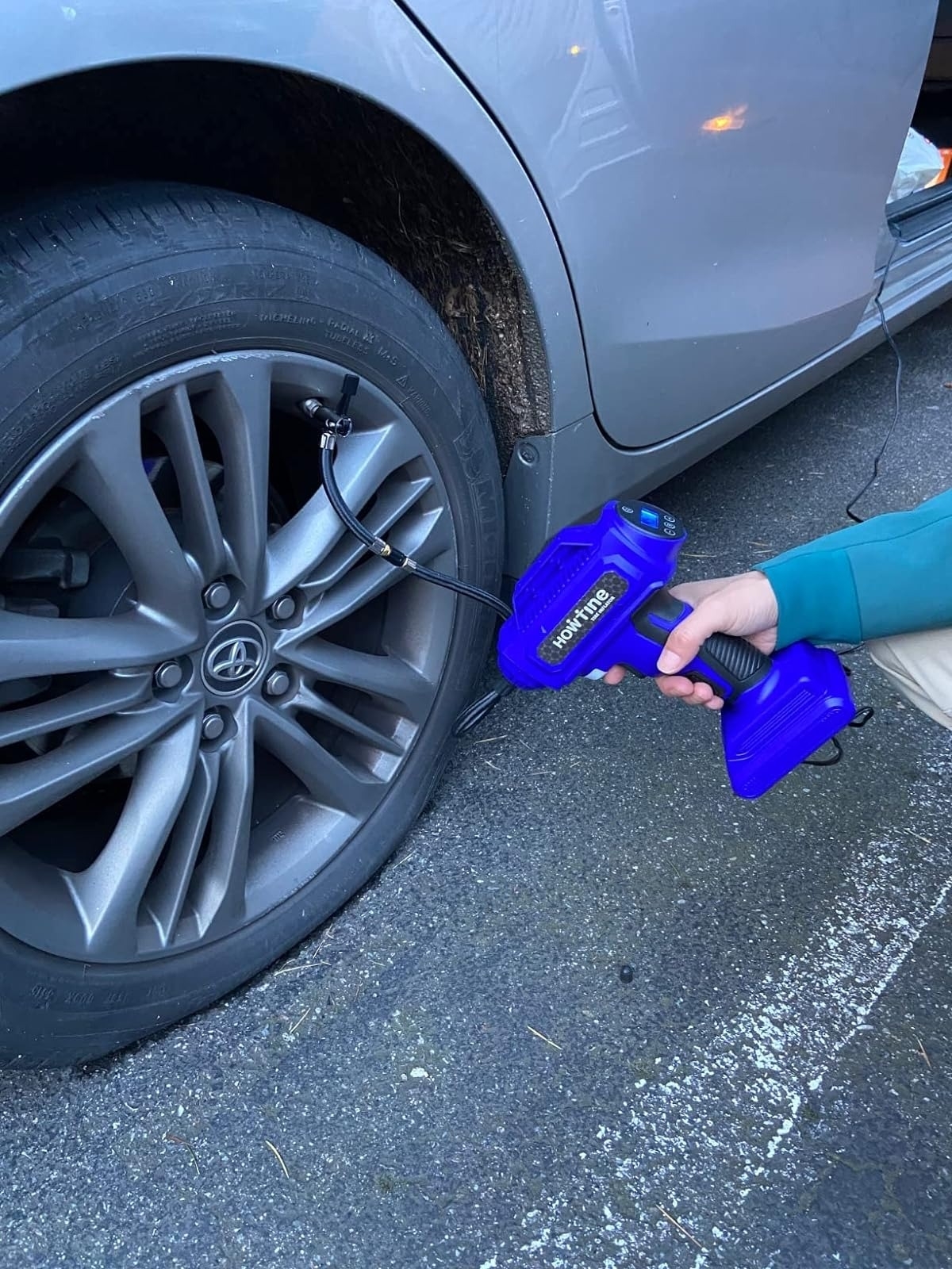 A person using a portable blue air compressor to inflate the tire of a car. The image is about a shopping article on useful automotive tools