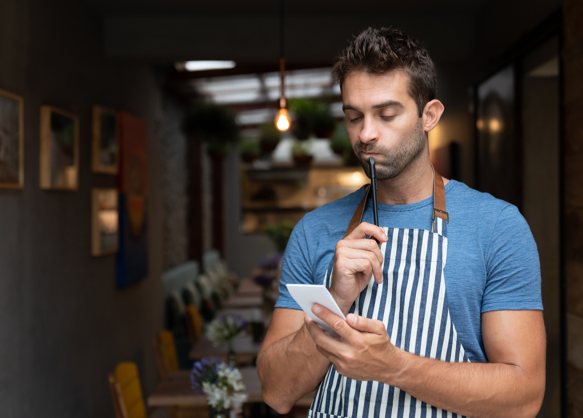 A man wearing a blue shirt and striped apron stands in a restaurant, holding a pen near his mouth and looking at a notepad