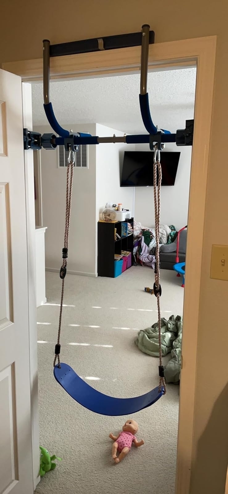 Door frame hanging swing in a child&#x27;s bedroom, with toys and storage shelves visible in the background. A baby doll lies on the carpet