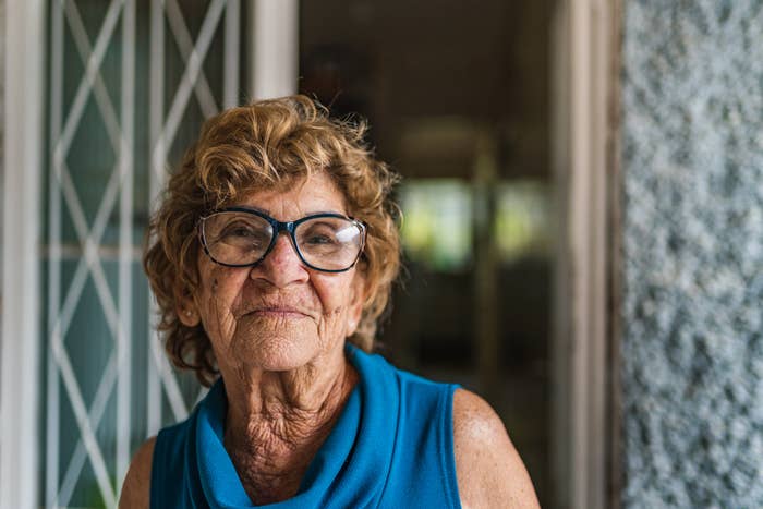 An elderly woman with curly hair and large glasses is smiling while standing in a doorway