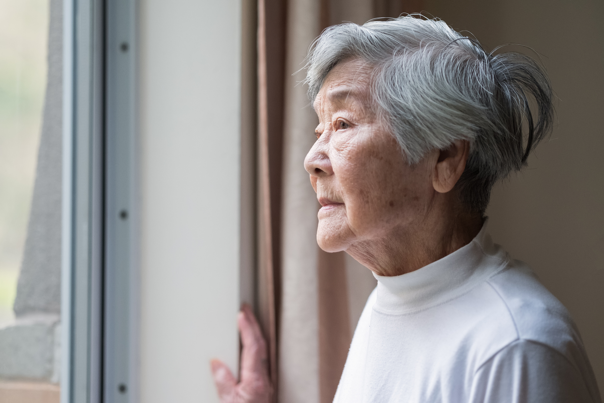 Elderly woman with short hair, in a white turtleneck, looking contemplatively out a window, hand resting on the curtain