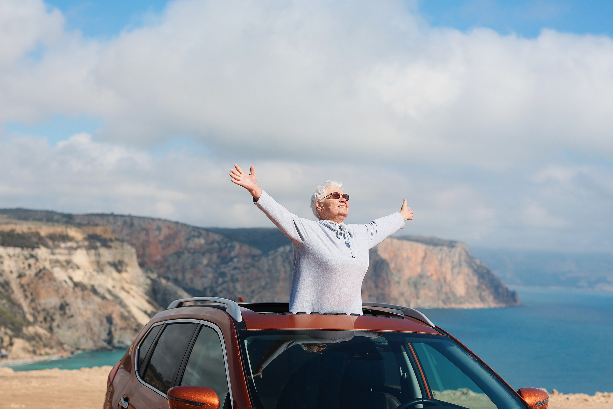 An elderly person stands through a car&#x27;s sunroof with arms outstretched, enjoying a scenic coastal view with cliffs and ocean in the background