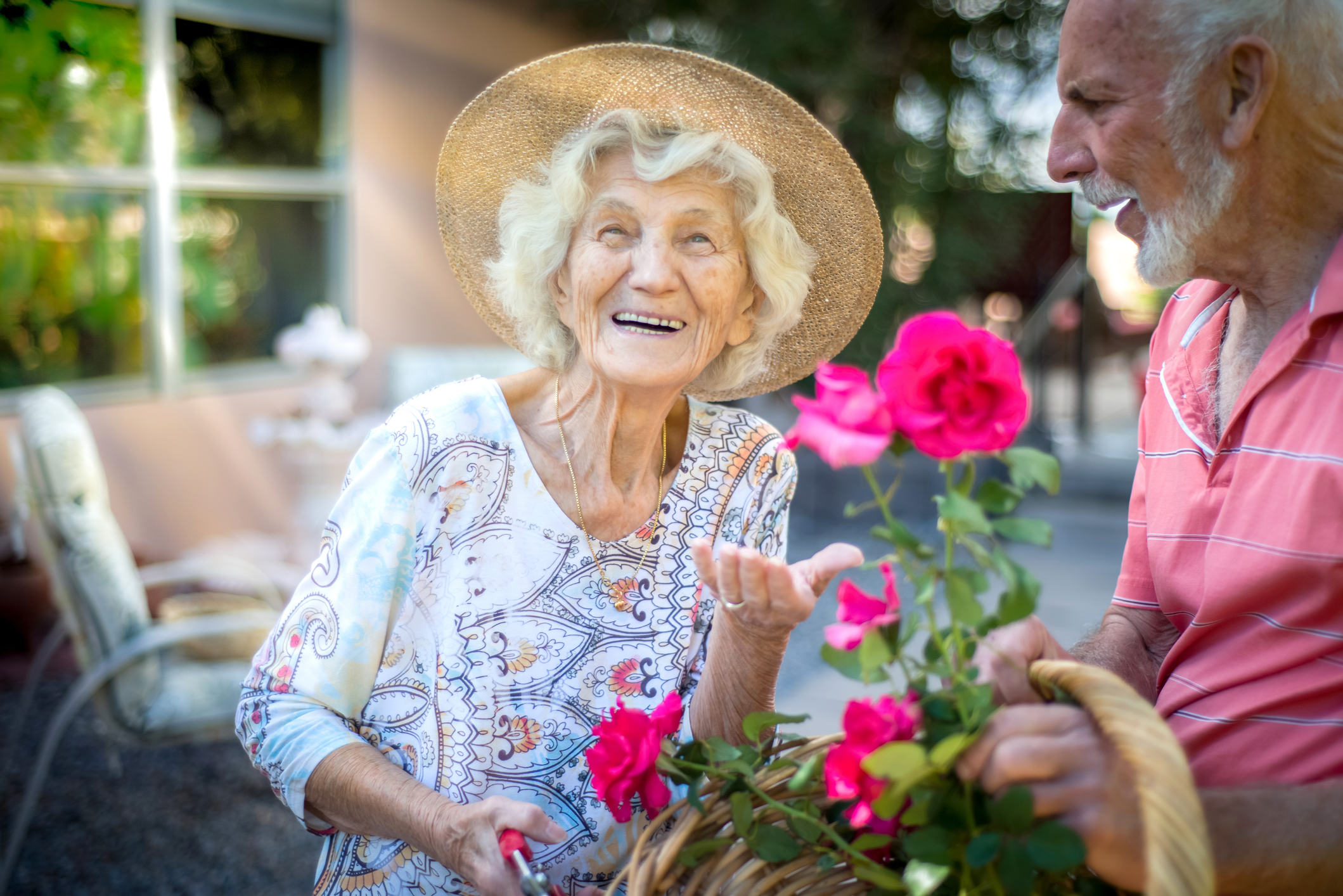 Elderly woman with a straw hat and paisley top happily tending to a rose garden with an elderly man in a casual shirt