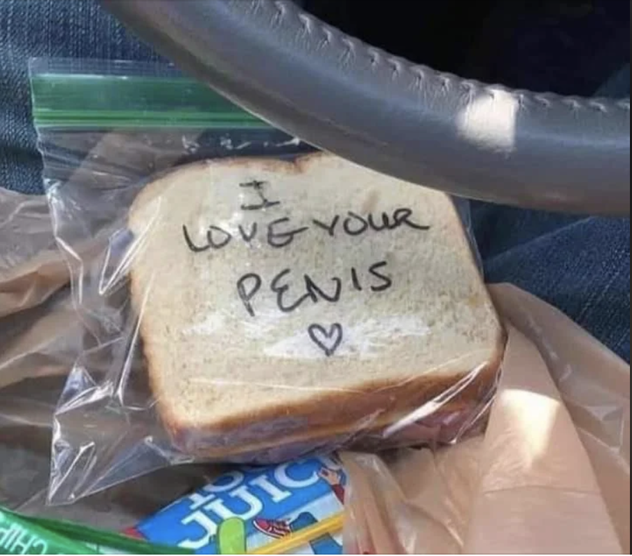 A sandwich in a plastic bag with text written on the bread: &quot;I love your penis&quot; and a heart symbol. The sandwich is on a car seat. Nearby, there&#x27;s a juice box