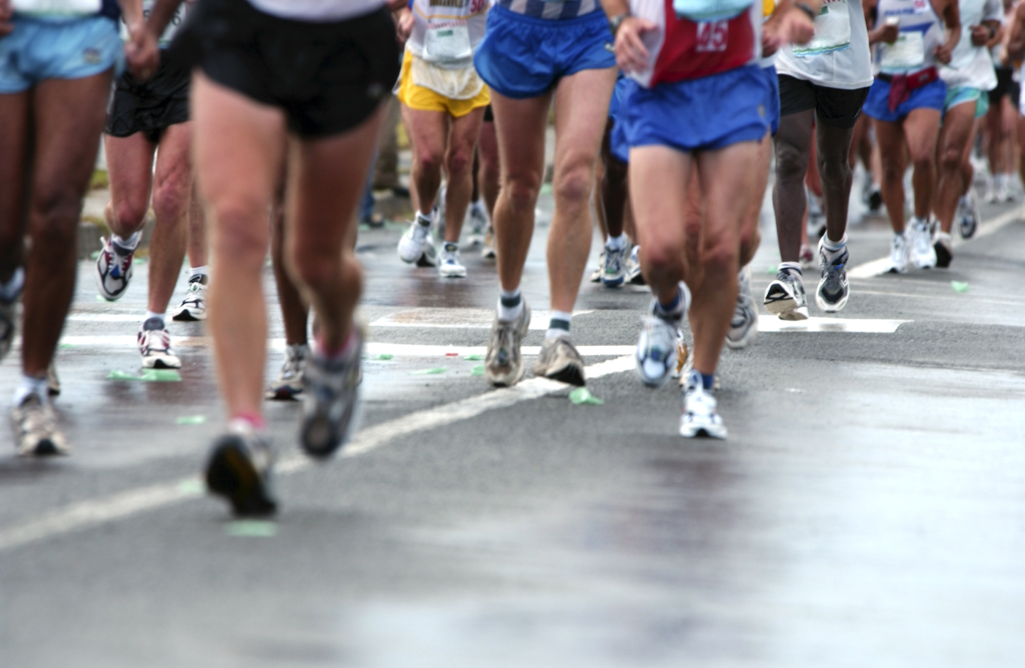 A group of marathon runners are seen running on a road. Their legs and running shoes are visible, but their upper bodies and faces are mostly out of frame