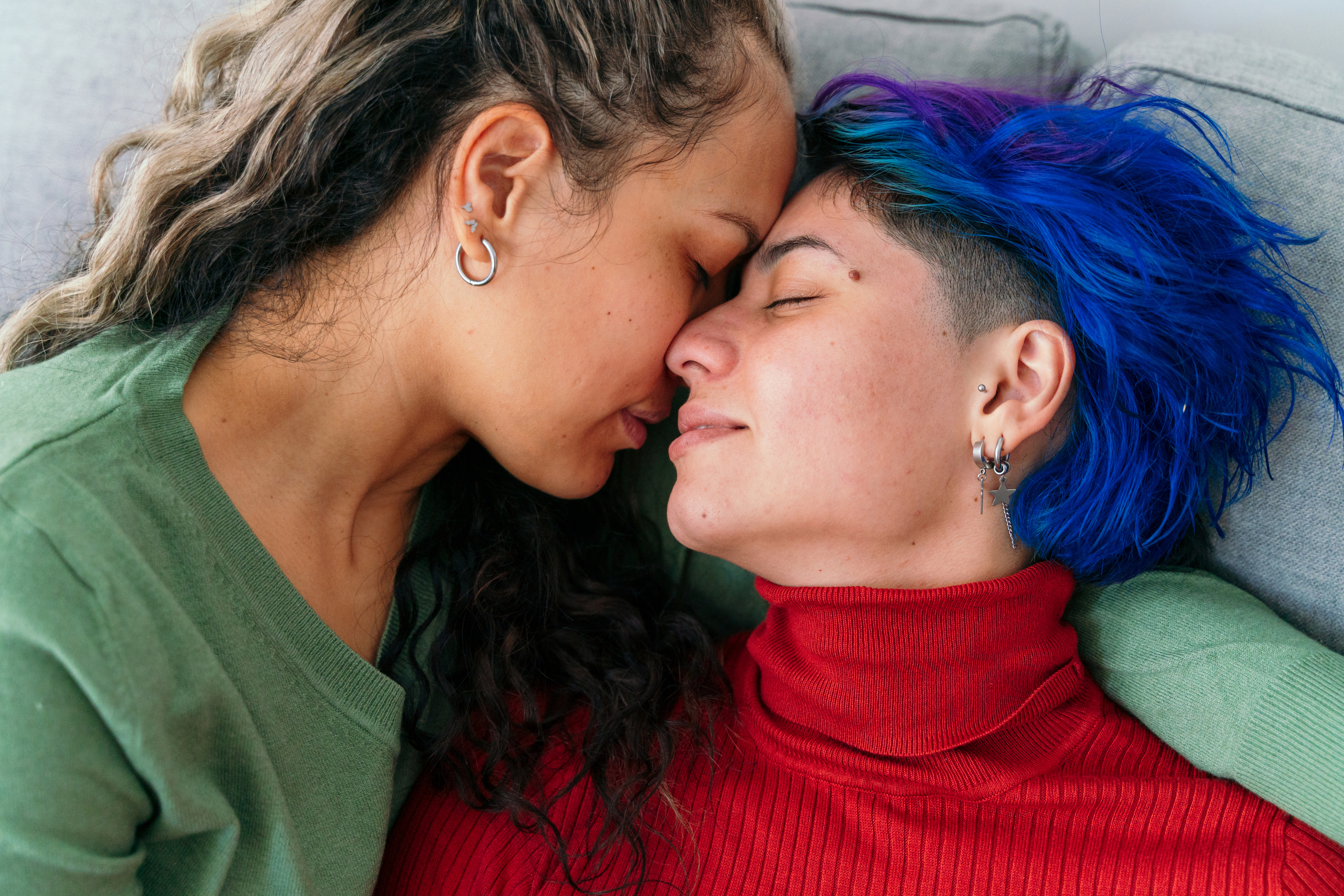 Two people, one in a green top and the other in a red turtleneck, share an intimate moment while snuggling on a couch