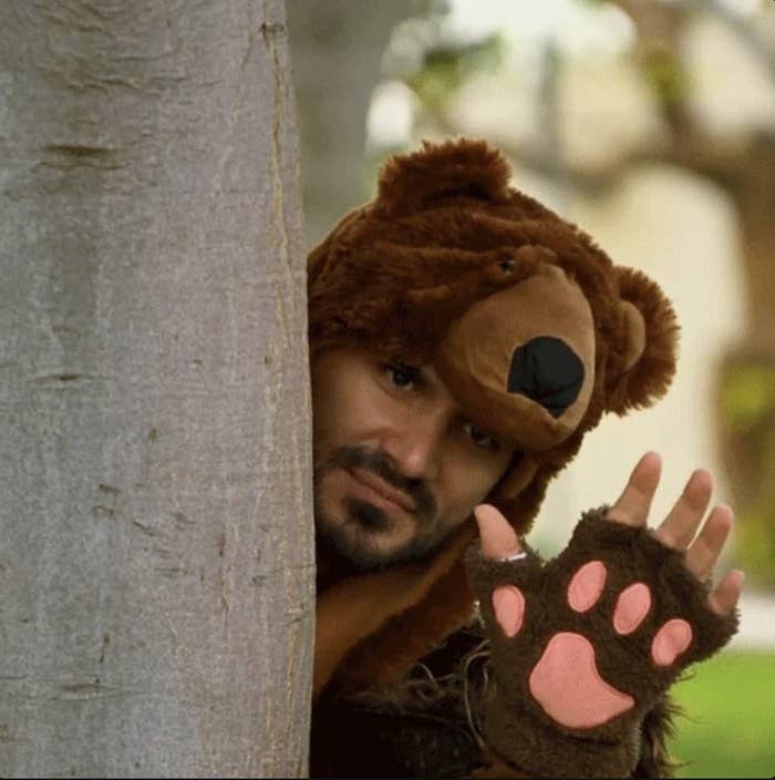 Man in a bear costume peeking from behind a tree and waving with one paw