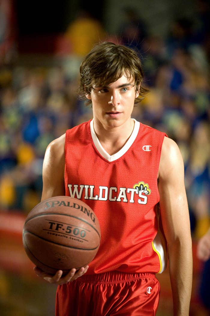 Zac Efron in a red Wildcats basketball jersey, holding a Spalding basketball, in a scene from High School Musical