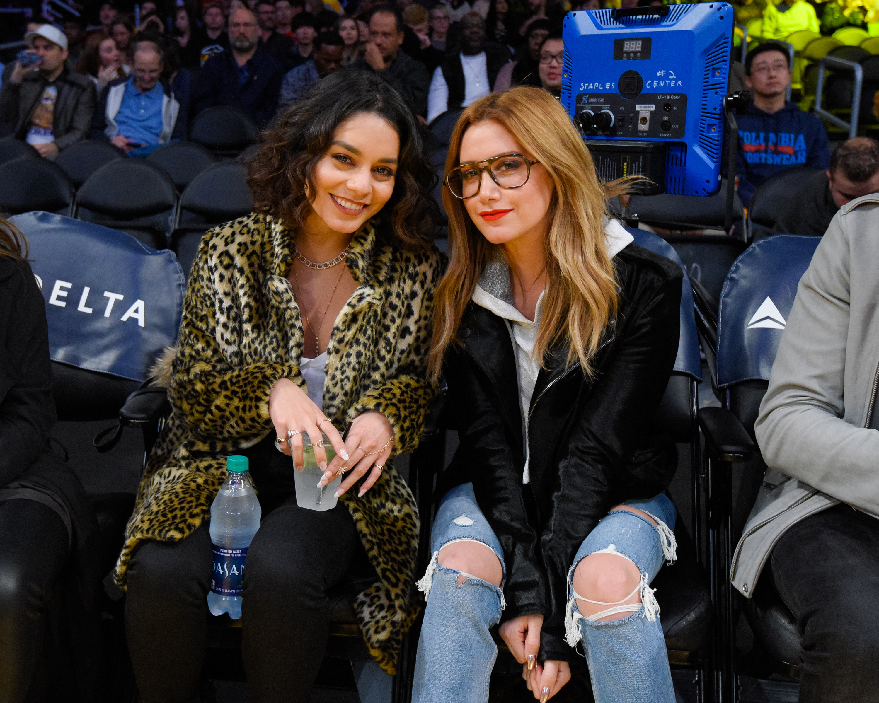 Vanessa Hudgens in leopard print coat and Ashley Tisdale in black jacket and ripped jeans sitting courtside at a basketball game