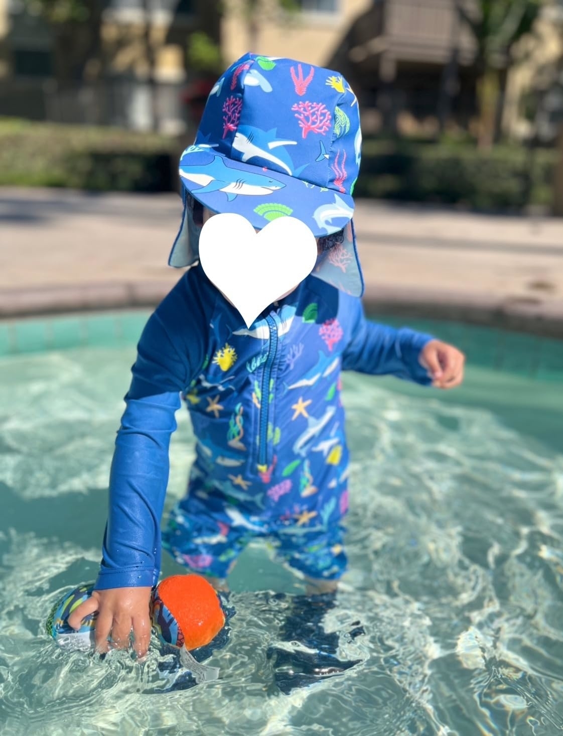 Reviewer&#x27;s photo of a toddler in a blue aquatic-themed zip-up swimsuit, playing with a toy in shallow water. The face of the child is covered with a white heart icon