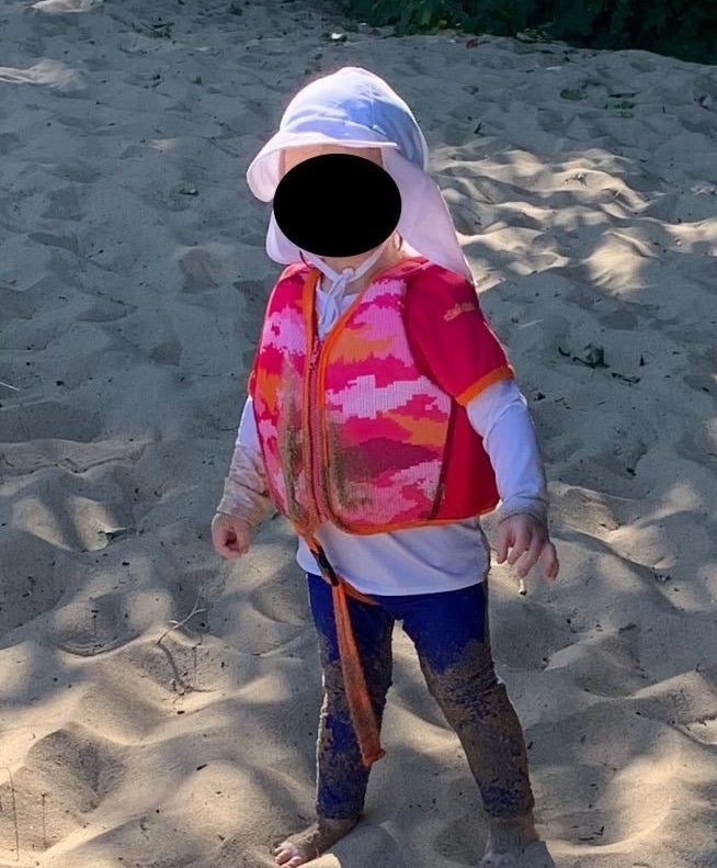 Reviewer&#x27;s photo of a toddler wearing a white sun hat, patterned life preserver vest, and rolled-up pants, standing barefoot on a sandy beach. The child&#x27;s face is obscured by a black circle