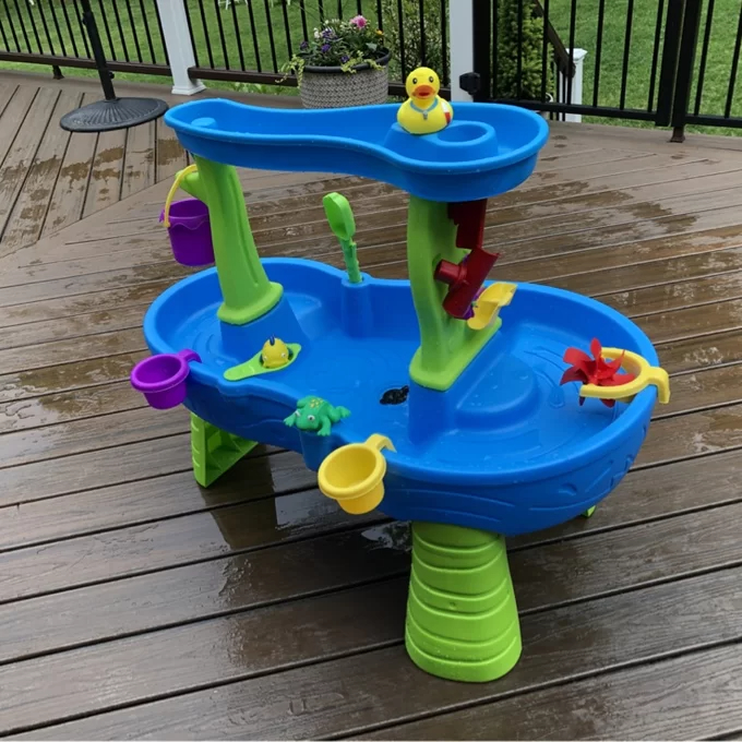 Reviewer&#x27;s photo of the water play table with various interactive toys, such as water wheels, buckets, and a rubber duck, set up on a wet wooden deck