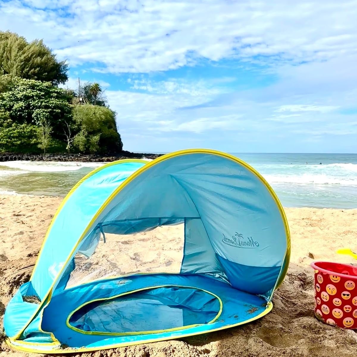 Reviewer&#x27;s photo of the pop-up beach tent set up on sandy beach with ocean and trees in the background. Toy bucket, cup, and shovel are in the foreground