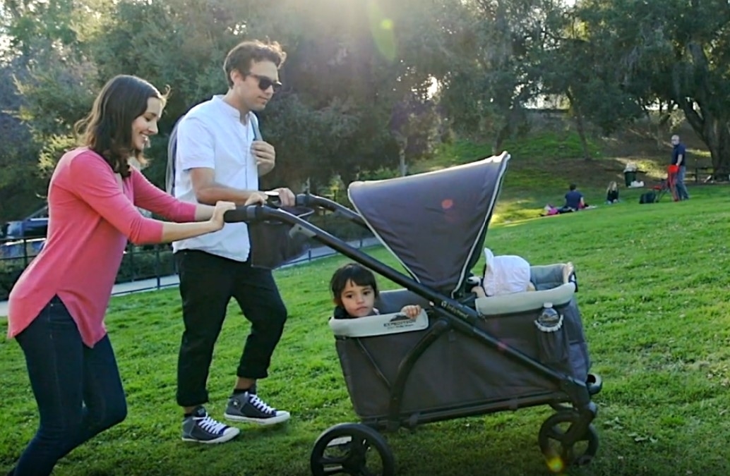 A parent pushes a wagon stroller with a child, while the other parent walks beside them in a park