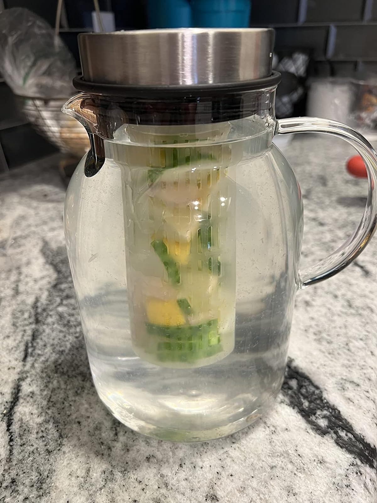 Glass pitcher filled with water and a filter insert containing cucumber and lemon slices on a kitchen countertop