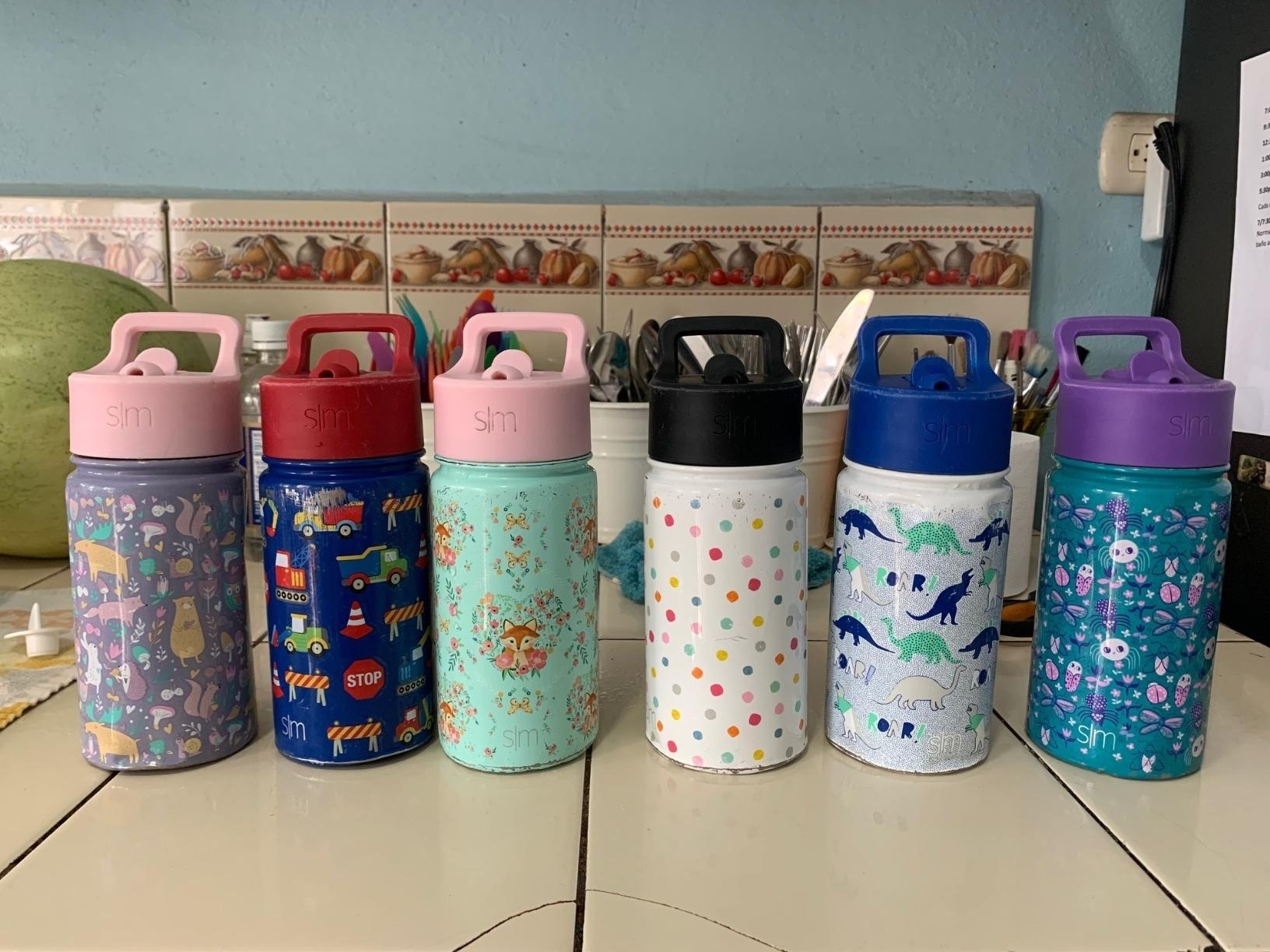 Six colorful water bottles with various patterns including animals, dinosaurs, polka dots, and abstract designs, displayed on a kitchen counter