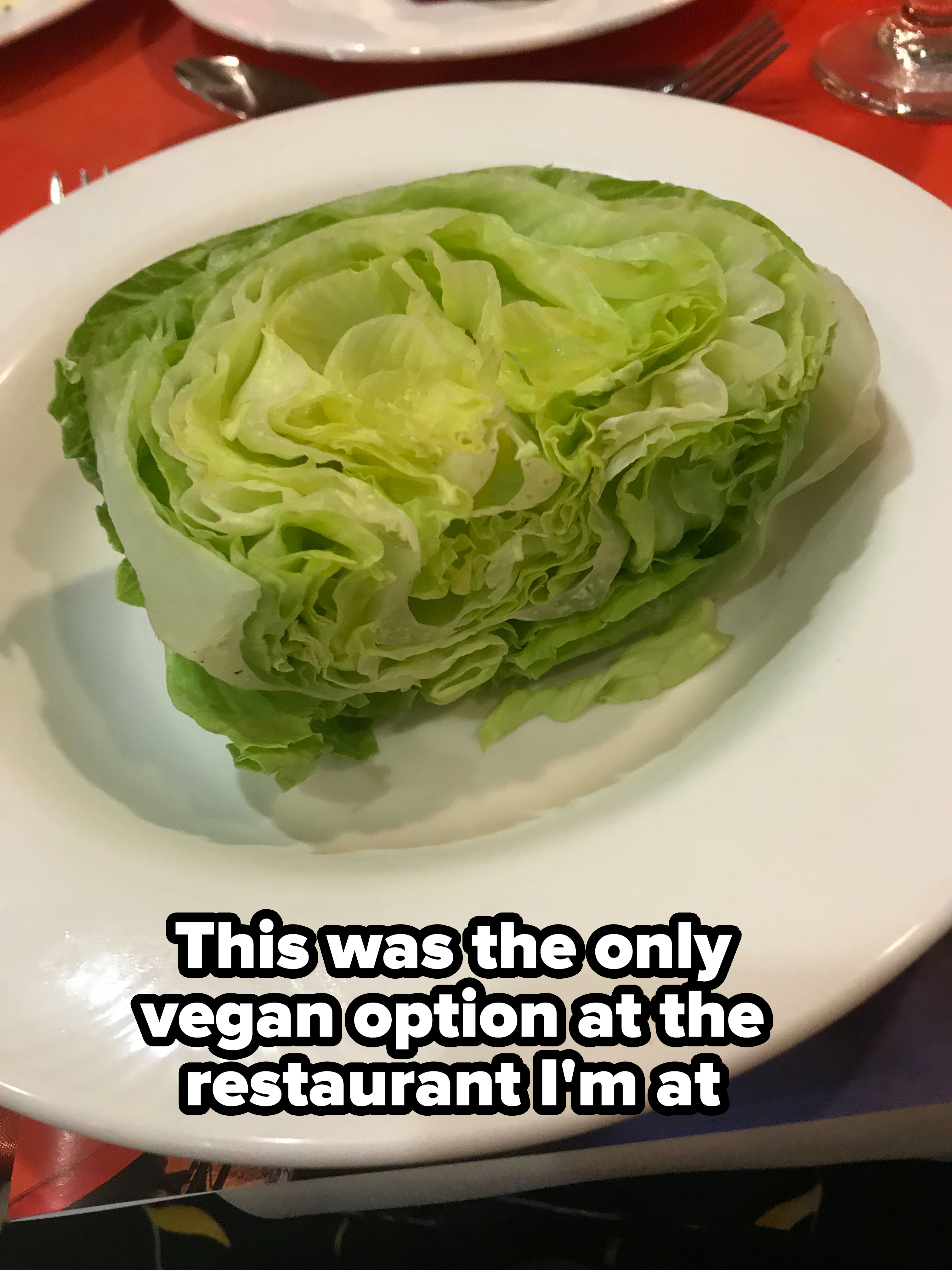 A wedge of iceberg lettuce is served on a white plate. The plate is placed on what appears to be a red tablecloth, with silverware and another plate in the background