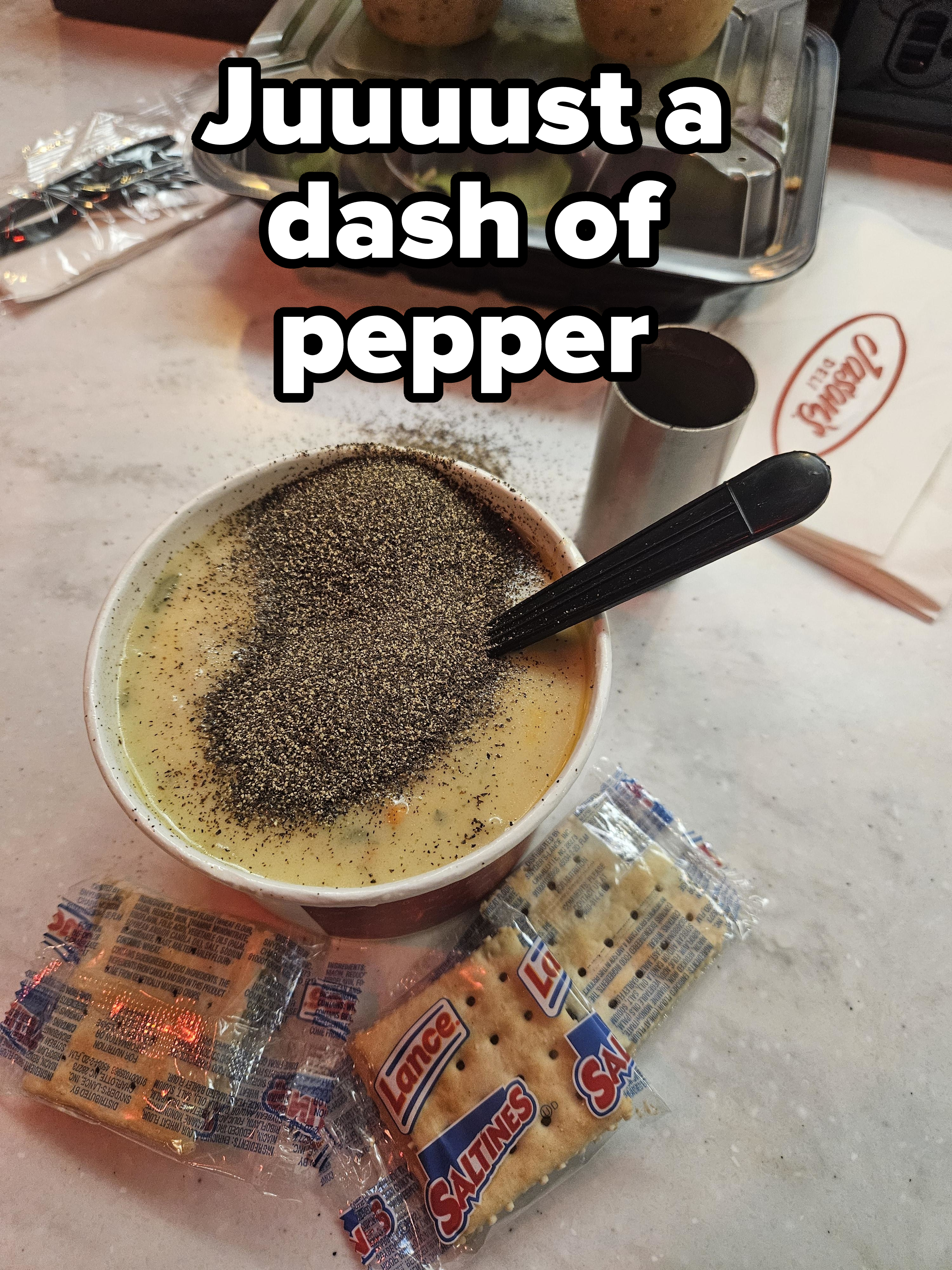 A bowl of soup heavily sprinkled with black pepper, a spoon, and packs of saltine crackers on a marble surface