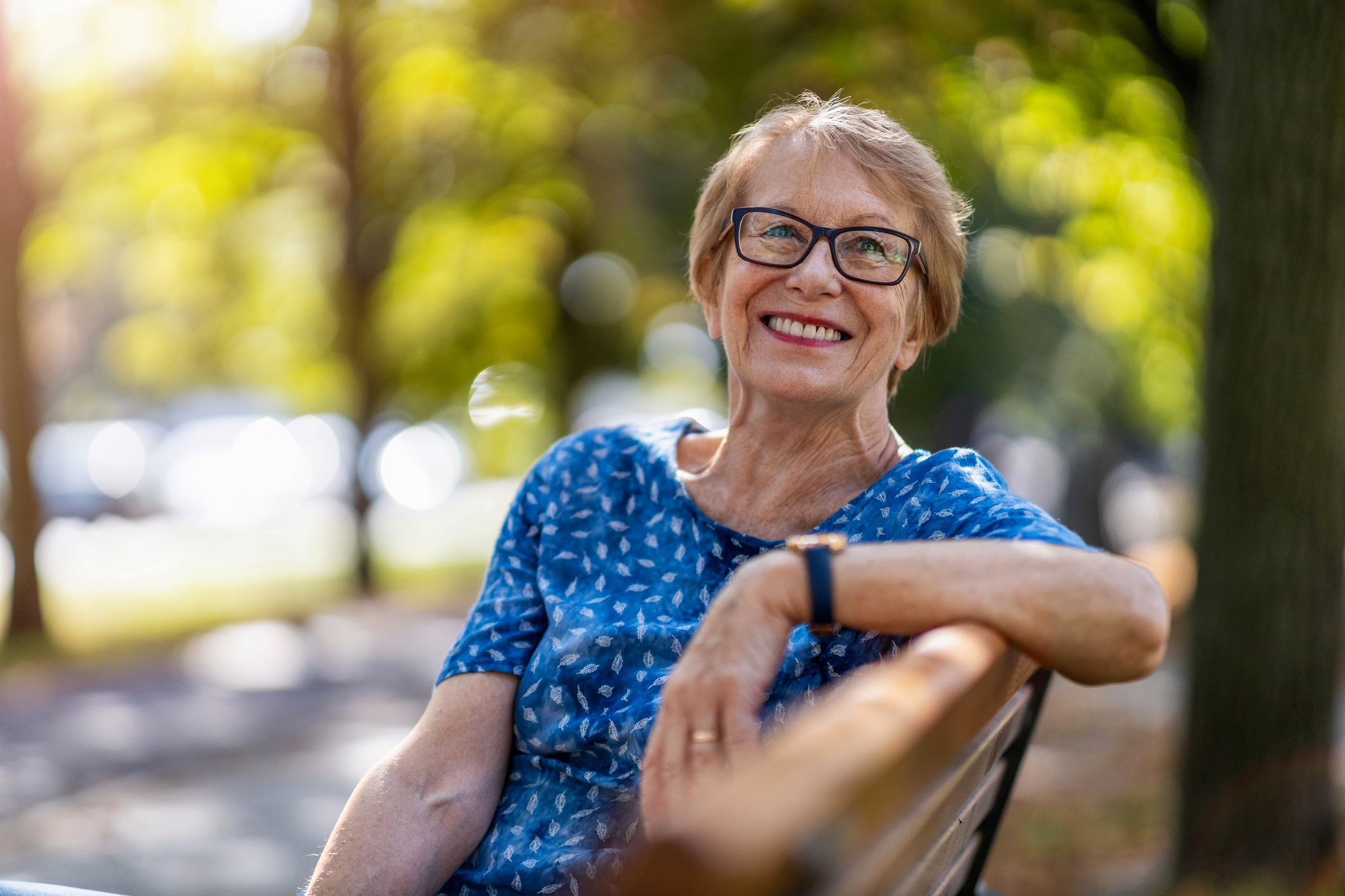 An older woman wearing glasses and a patterned blouse sits on a park bench, smiling at the camera. Trees and sunlight are visible in the background