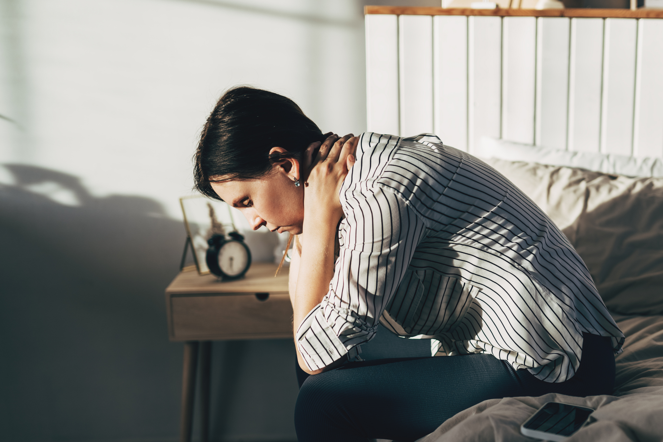 A person with short hair sits on a bed holding their neck, in a pensive or painful posture. An alarm clock and phone are on the bedside table