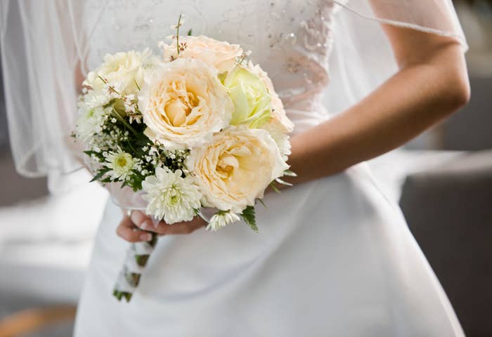 A bride holds a bouquet of large roses and delicate flowers, wearing a white wedding dress with intricate beadwork, and a veil draped over her shoulders