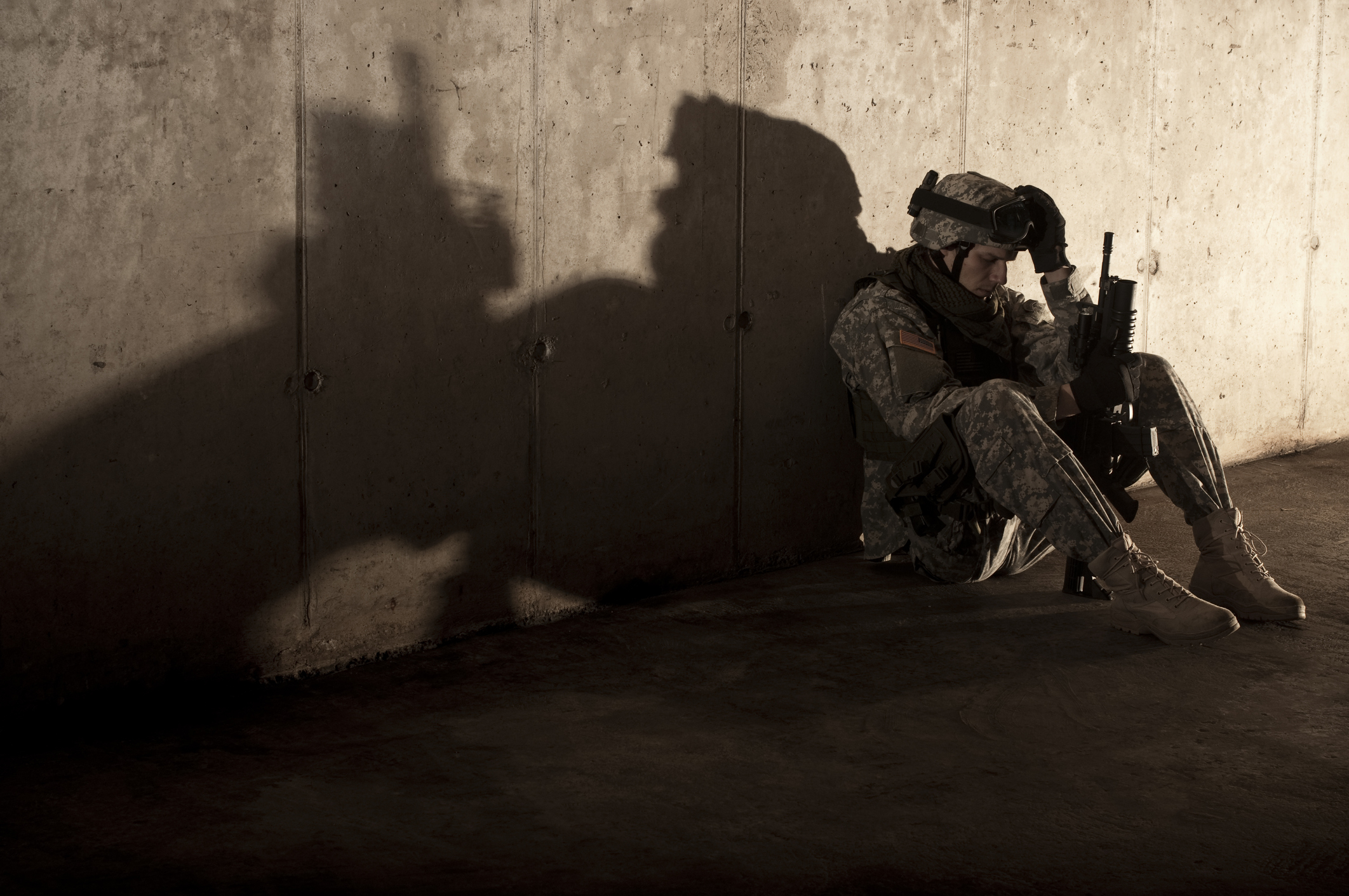 A soldier in military gear sits on the ground, head down, holding a rifle. The shadow on the wall shows the outline of Batman