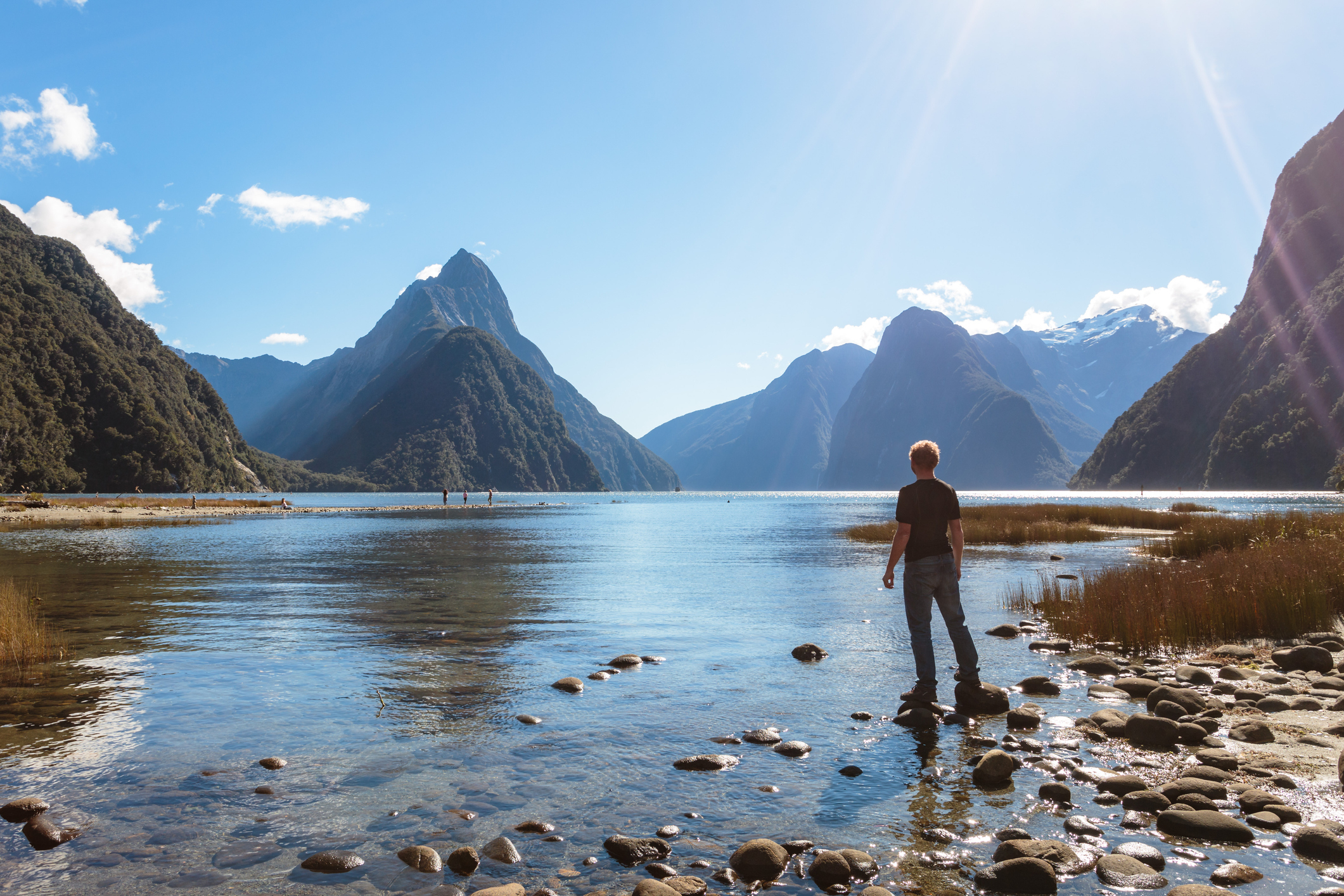 A person stands on rocky shore looking at a serene lake with mountains in the background. Sunlight casts a warm glow over the scene