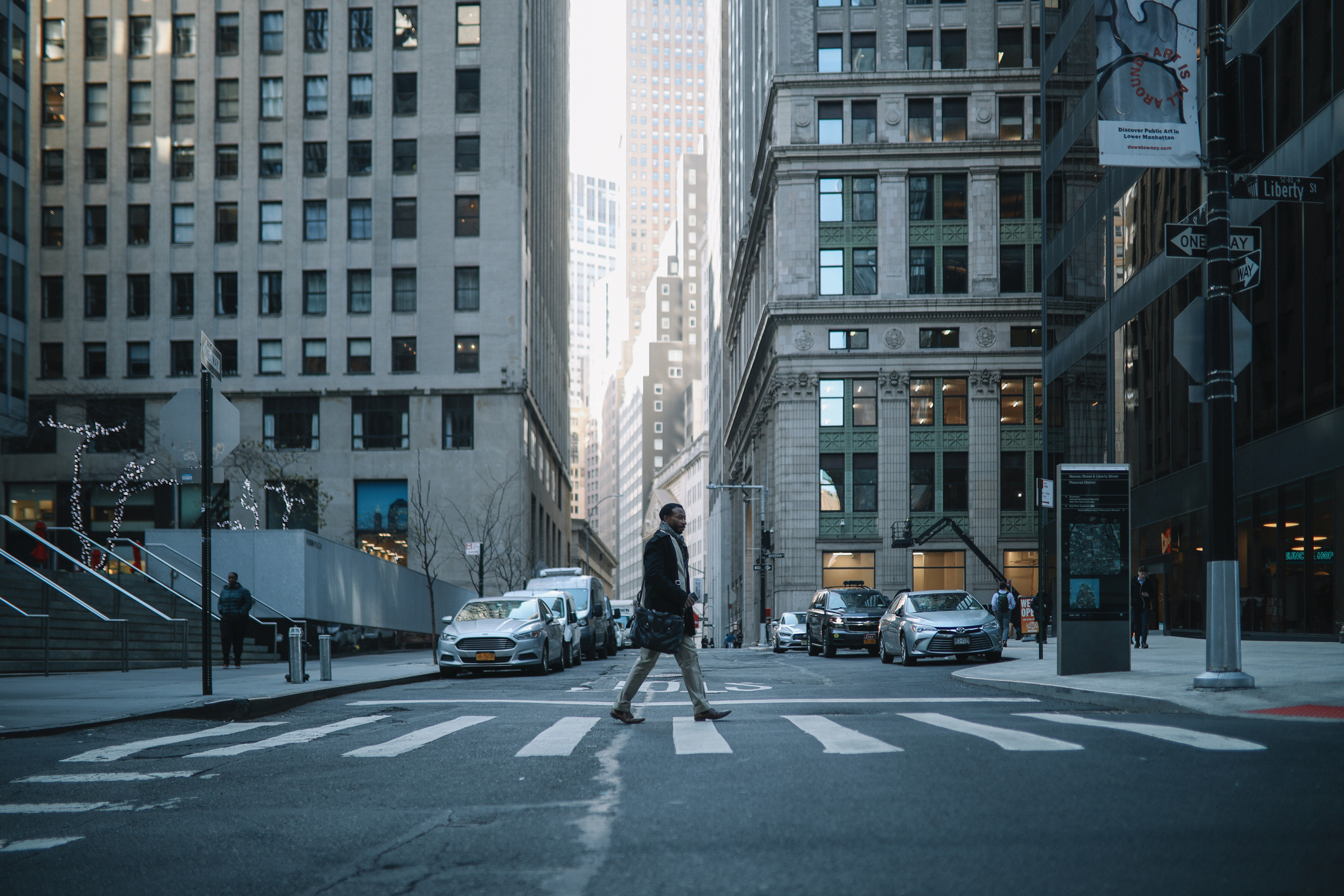 A person crosses a city street at a crosswalk, surrounded by tall buildings and parked cars