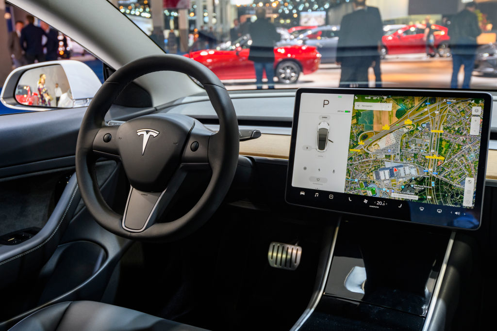 Tesla car interior showing the steering wheel with the Tesla logo, a touchscreen displaying navigation, and part of the car&#x27;s sleek dashboard design