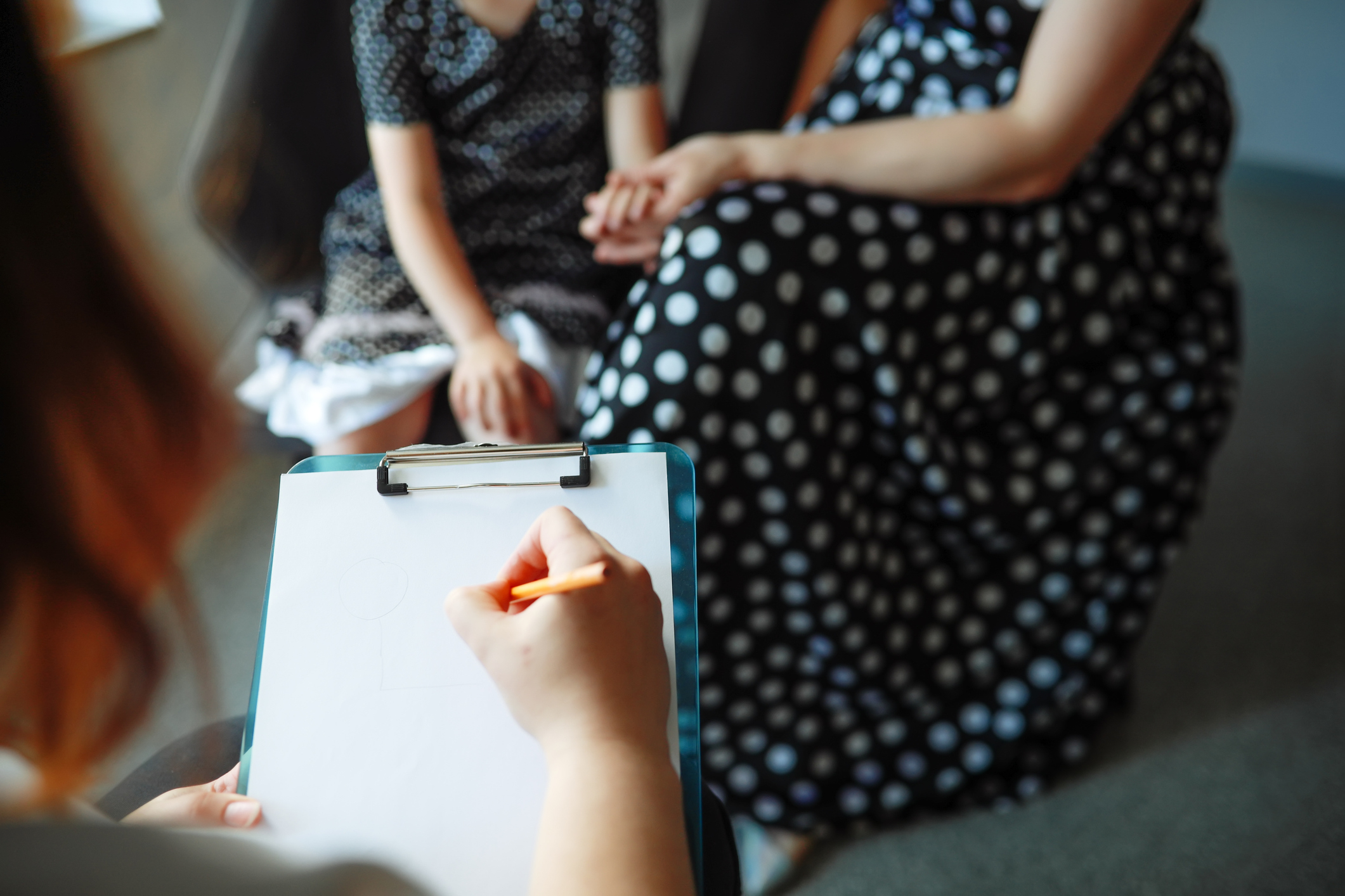 A woman holding a clipboard writes notes while two people, one adult and one child, both wearing polka dot dresses, sit and hold hands in the background