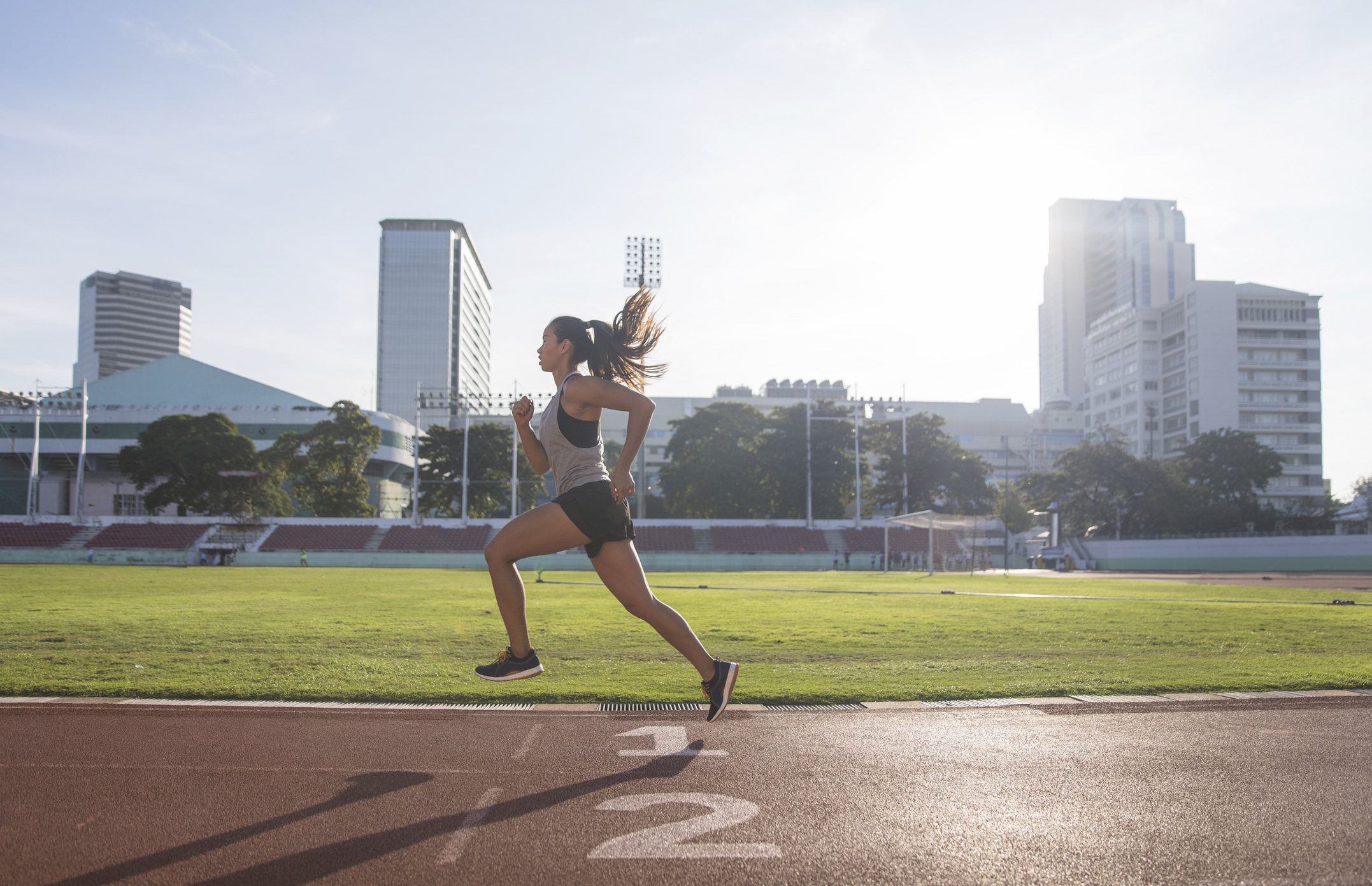 A woman is running on a track in an urban stadium with skyscrapers and greenery in the background
