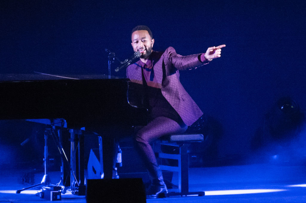 John Legend performs at a piano on stage, wearing a stylish suit, and pointing outward
