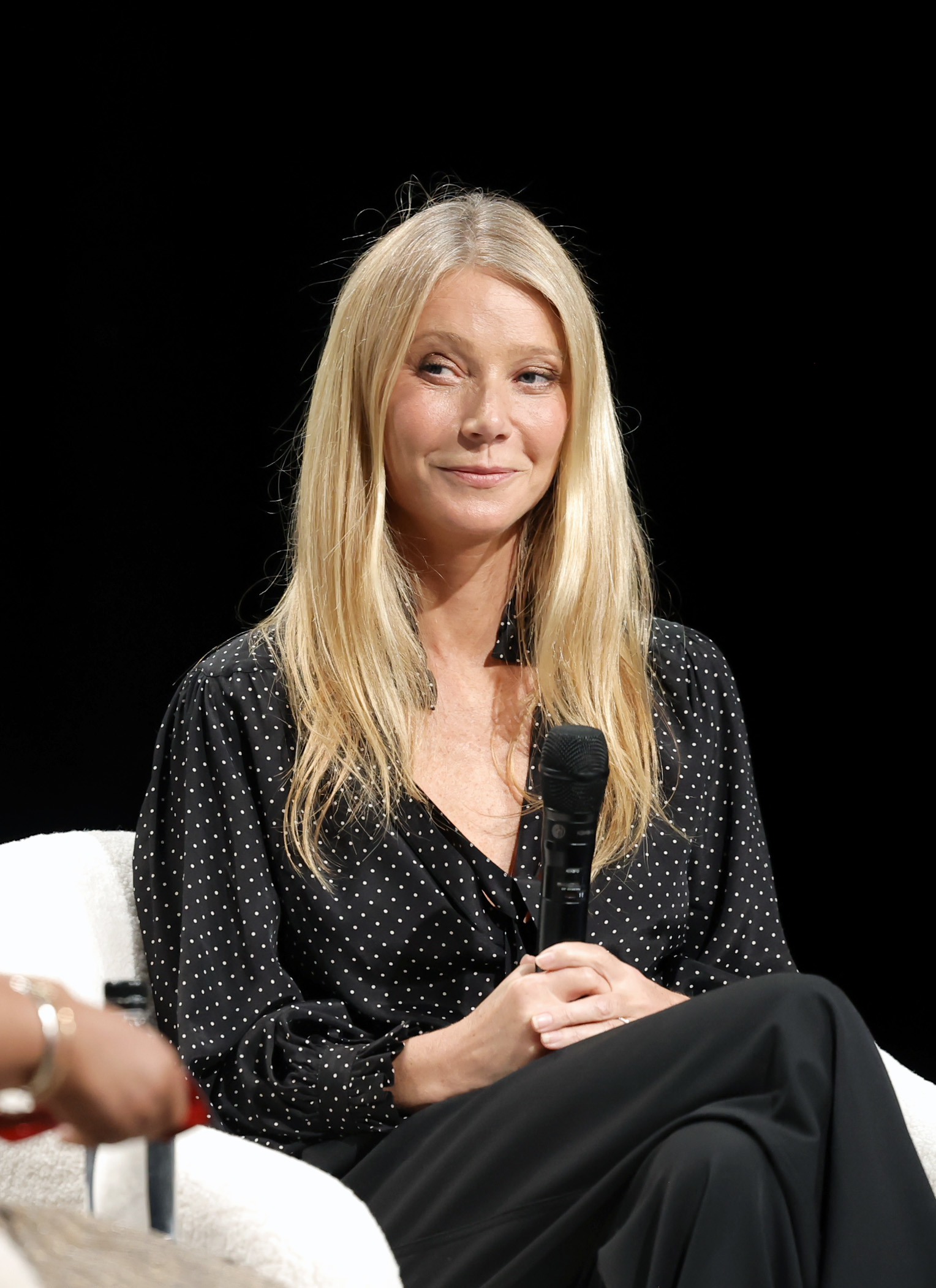 Gwyneth Paltrow sitting with a microphone, wearing a black polka-dot blouse and black pants, smiling during a discussion event