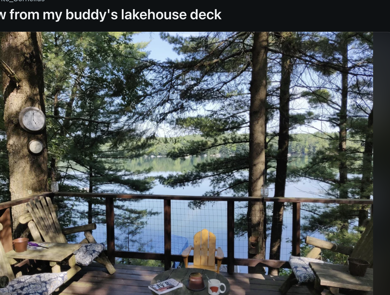 A scenic view from a wooden deck overlooking a lake surrounded by trees, with Adirondack chairs and a small table