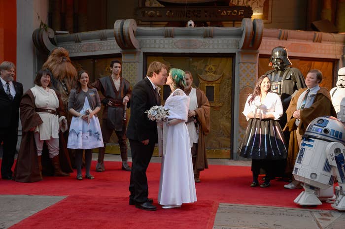 A wedding couple kisses on a red carpet surrounded by people in Star Wars costumes, including Darth Vader, R2-D2, and Chewbacca