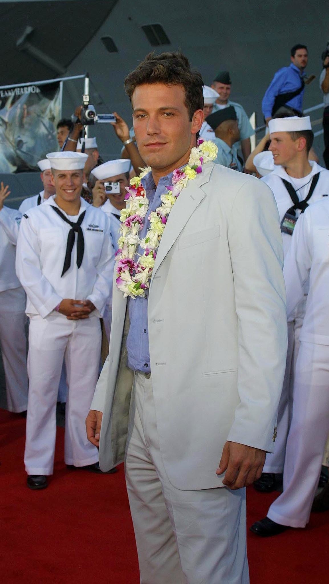 Ben Affleck wearing a lei and a light-colored suit on a red carpet, surrounded by smiling Navy personnel in uniforms