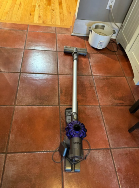 Dyson vacuum cleaner placed on a tiled kitchen floor near a wall outlet with cord attached. Wooden flooring seen in the background