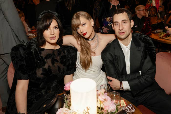 Lana Del Rey, Taylor Swift, and Jack Antonoff sit together at a formal event, smiling at the camera. They are dressed in elegant evening attire