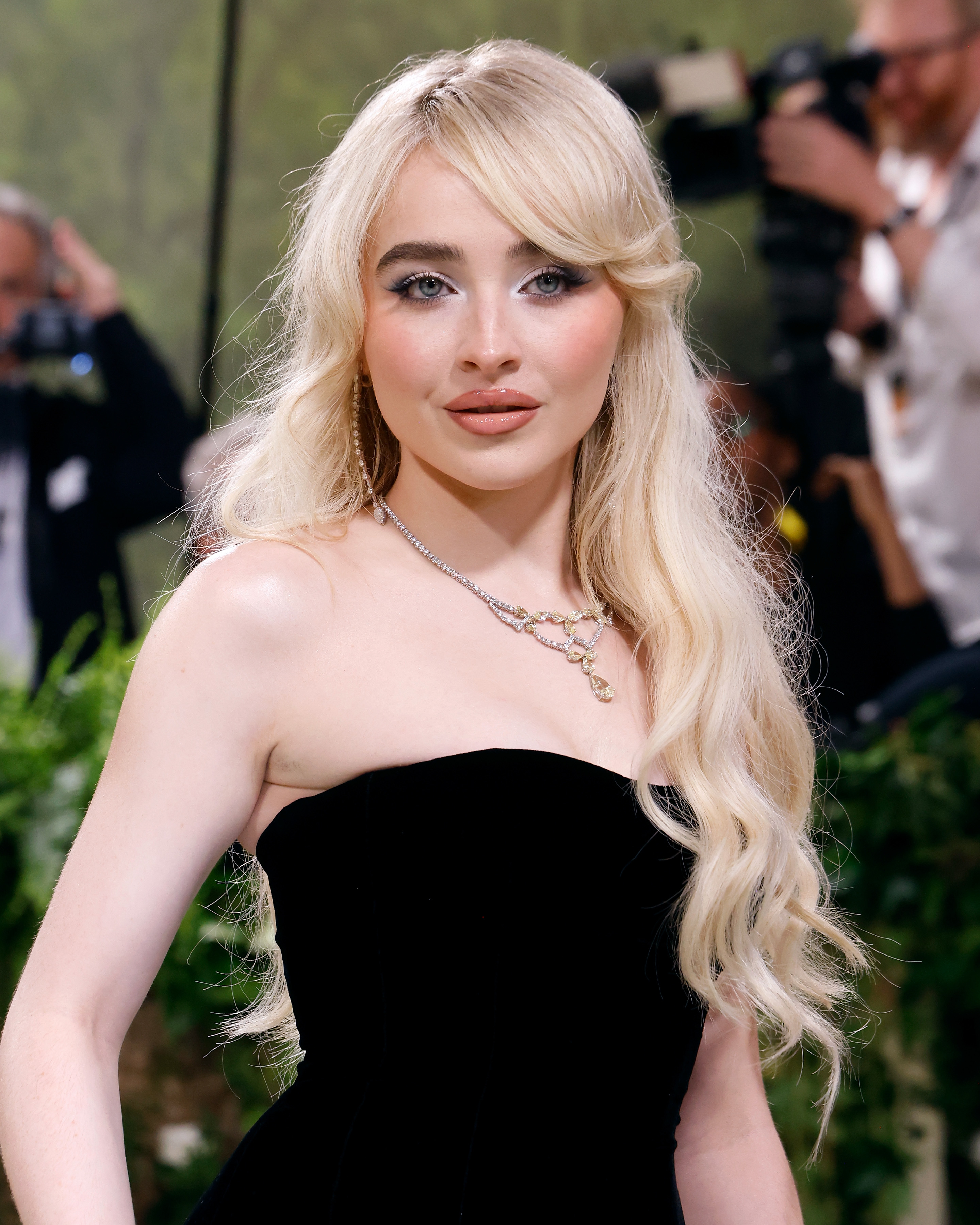 Sabrina Carpenter poses on the red carpet in a strapless, fitted dress, with wavy long hair, accessorized with a necklace and light makeup. Photographers are in the background