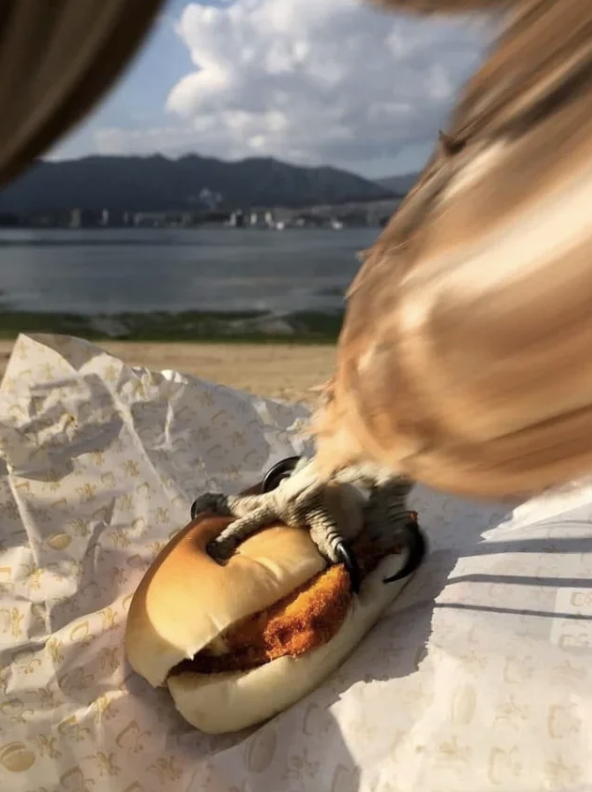 A bird is grabbing a hot dog from a bun, with a scenic lake and mountain background in the distance